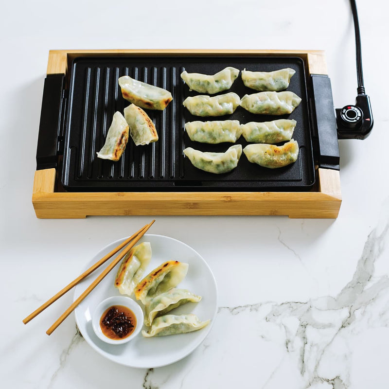 Dumplings on an electric grill and a plate with chopsticks and dipping sauce.