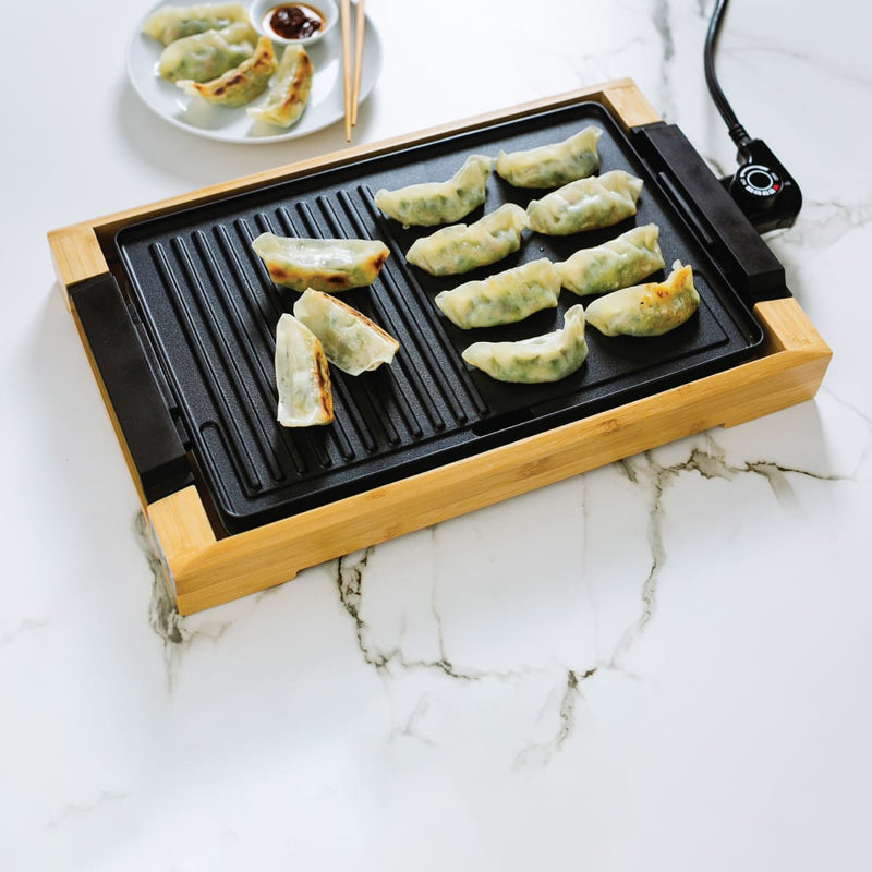 Dumplings on a black electric grill with a plate of soy sauce on a marble table.