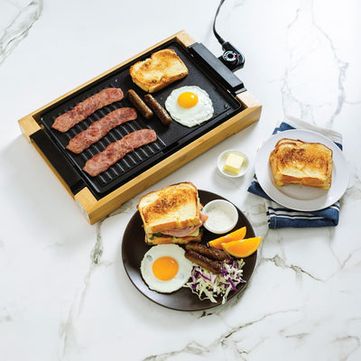 Electric griddle cooking bacon, sausages, egg, and toast, with a plated breakfast beside it.